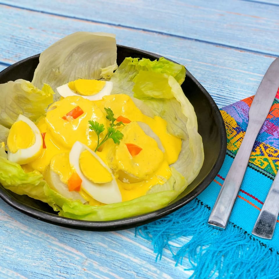 Peruvian Papa a la Huancaína with yellow sauce, hard-boiled eggs, and lettuce on a blue wooden table.