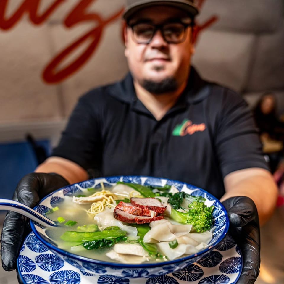 Chef in glasses and black gloves presenting a bowl of Peruvian wonton soup with pork and vegetables.