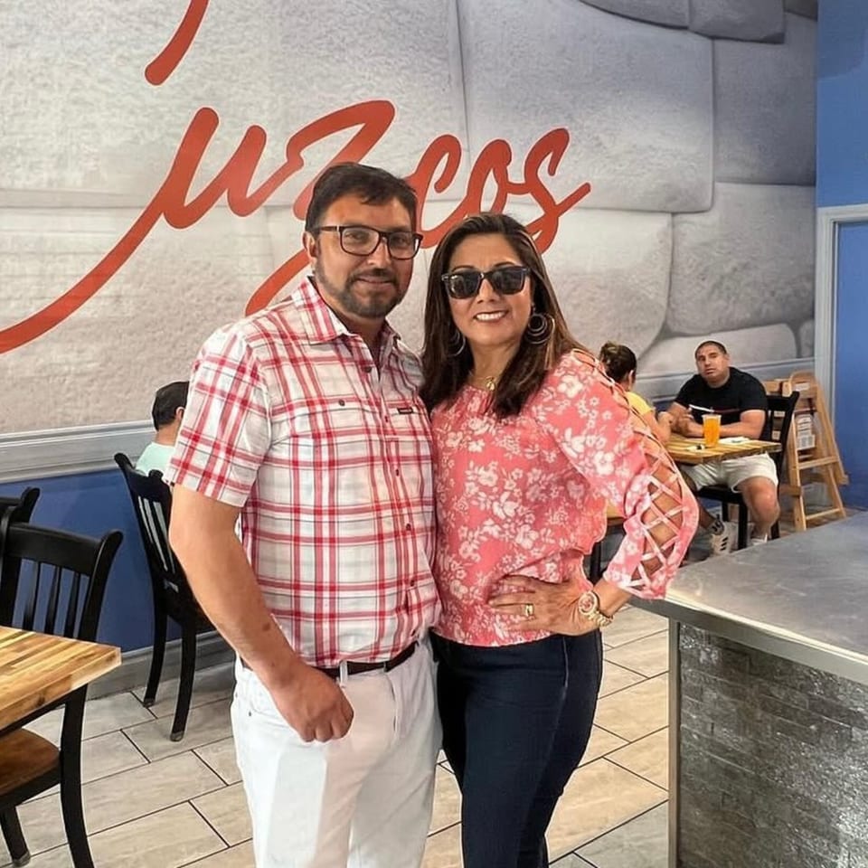 A couple smiling in front of a Cuzco's Peruvian Restaurant wall with the restaurant's name in red.