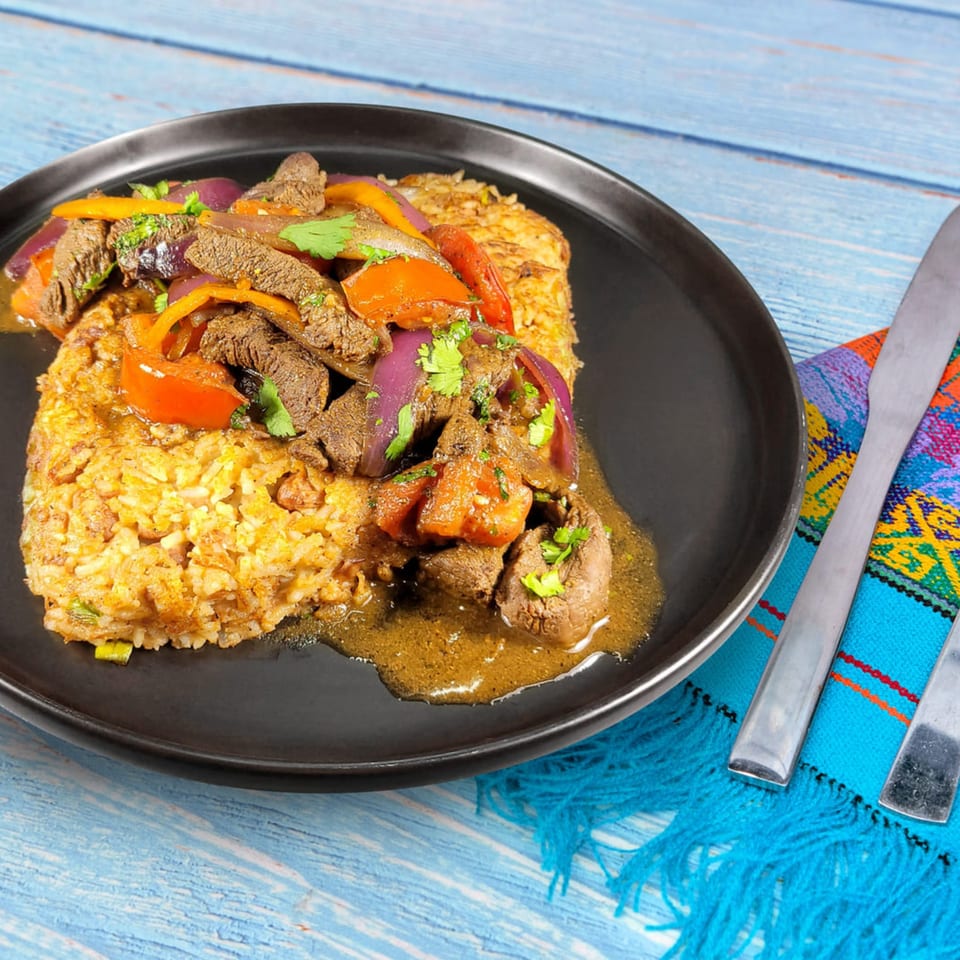 Peruvian Lomo Saltado with beef, red onions, tomatoes, and rice on a black plate.