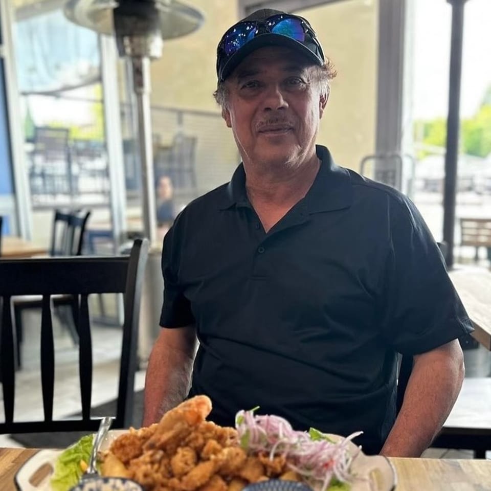 Man in a black polo shirt and cap sitting at a table with a plate of Peruvian fried seafood.